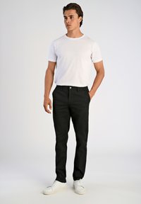 Hombre de pie vestido con una camiseta blanca de manga corta, pantalones negros con las manos en los bolsillos y zapatillas blancas, frente a un fondo claro y liso.