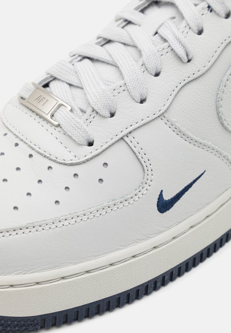 Białe skórzane sneakersy z perforowanym przodem, teksturowanymi akcentami i granatowym logo Nike. Posiadają pasujące sznurowadła i gumową podeszwę.