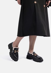 Mocassini in pelle nera con un rivestimento beige morbido, dettagli in metallo dorato e suole in gomma spesse, progettati per il comfort e la durata.