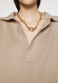 Close-up van een persoon die een chunky asymmetrische gouden ketting draagt en een beige coltrui met V-hals.