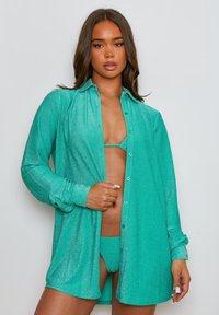 Camicia oversized turchese scintillante con chiusura a bottoni, maniche lunghe e colletto, indossata sopra un bikini abbinato. Texture liscia.