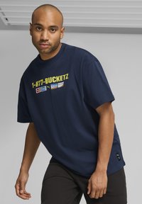 Zilā kokvilnas T-krekls ar īsām piedurknēm, ar dzeltenu tekstu "1-877-BUCKETZ" un logotipiem. Melni bikses ar mazu zīmola birku uz apakšējās malas.