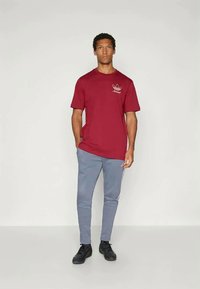 Burgundi színű pamut t-shirt rövid ujjakkal és kicsi fehér logóval a mellrészen, világosszürke, oldalsó csíkos, tapered fazonú melegítőnadrággal párosítva.