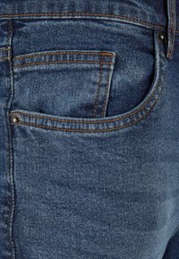 Denim-kangas tummansinisellä sävyllä, jossa on hieman haalistunut rakenne. Sisältää takataskun, jossa on näkyvät saumat ja metallipainike.