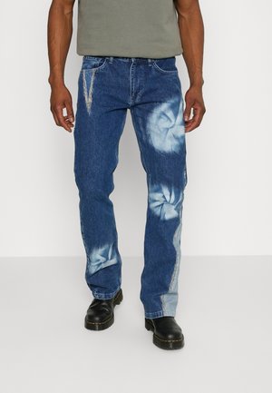 Man die een blauwe spijkerbroek draagt met gebleekte tie-dye patronen en zwarte laarzen, staand tegen een effen lichte achtergrond.