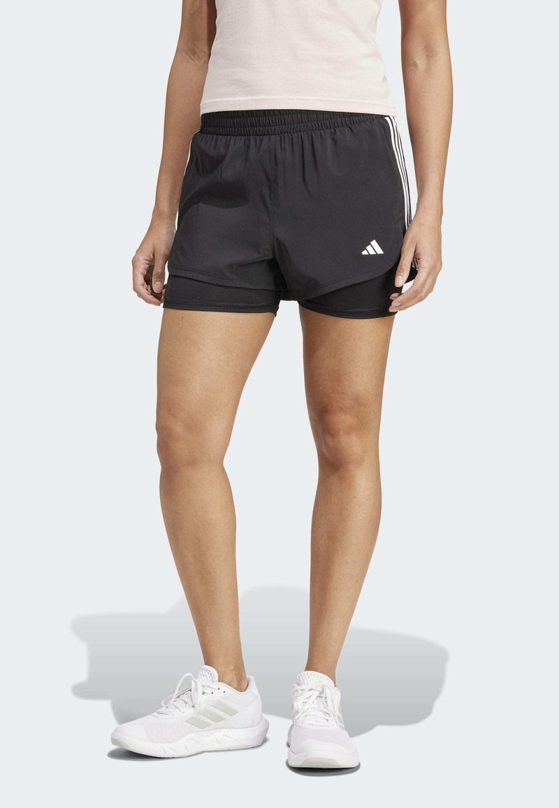 Femme portant un short de sport noir superposé avec des bandes blanches et des baskets blanches, debout devant un fond uni clair.