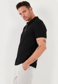 Fekete polo ing, cipzáras gallérral, rövid ujjú, és textúrált anyagból készült. Világos színű nadrággal viselve, látszanak a karokra tetoválások.