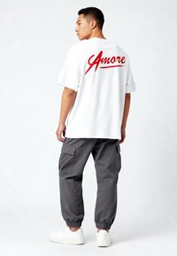 Witte katoenen t-shirt met rode 'Amore' tekst op de achterkant, gecombineerd met grijze cargobroek en witte sneakers. Relaxte pasvorm, korte mouwen.