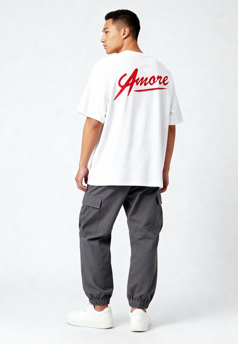 Witte katoenen t-shirt met rode 'Amore' tekst op de achterkant, gecombineerd met grijze cargobroek en witte sneakers. Relaxte pasvorm, korte mouwen.