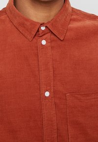 Camisa de pana color óxido con collar abotonado, botones blancos y un bolsillo parche en el lado izquierdo. Tejido ribeteado texturizado.