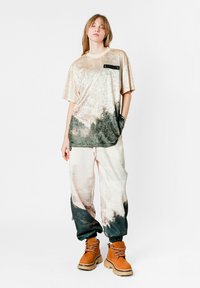 Camiseta de terciopelo en beige claro con degradado verde oscuro; cuenta con un bolsillo negro. Combínala con pantalones blancos texturizados y botas marrón claro.