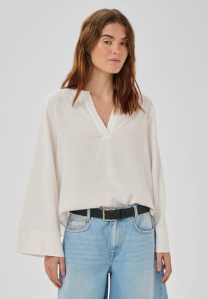 Kvinde med skulderlangt brunt hår iført en løs hvid V-hals-bluse stukket ned i lyseblå jeans med sort bælte.