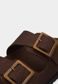 Καφέ σουέντ σανδάλι με δύο ρυθμιζόμενους ιμάντες που διαθέτουν ορθογώνιες μπρονζέ αγκράφες, σε λευκό φόντο.