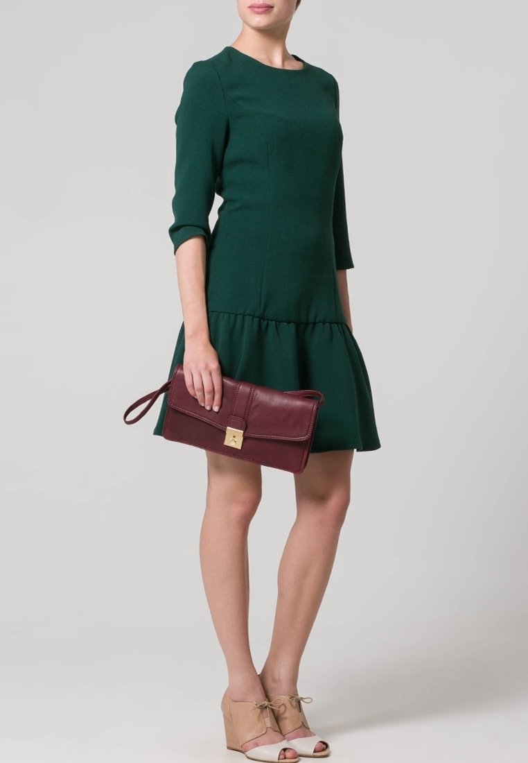 Femme portant une robe vert foncé à manches trois-quarts, tenant une pochette bordeaux et portant des talons compensés beiges.