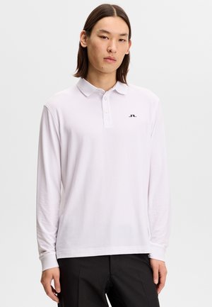 Młody mężczyzna z prostymi włosami, ubrany w białą koszulkę polo z długim rękawem i czarne spodnie, stojący na tle jednolitego, jasnego tła.