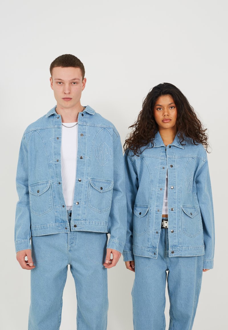 Jachete din denim de culoare bleu deschis, cu guler ascuțit, buzunare frontale și butoane argintii. Ambele modele poartă pantaloni din denim albi în nuanța bleu.
