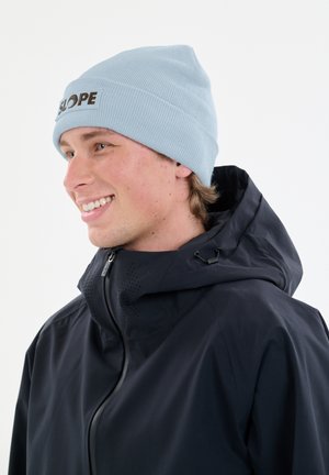 Lichtblauwe gebreide muts met een omgeslagen rand waar het woord "SLOPE" in het bruin op staat. Draagt bij een zwarte waterdichte jas met een ritssluiting.