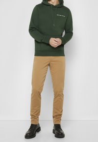 Dark green hoodie with a kangaroo pocket and drawstrings, paired with khaki pants and black lace-up boots. Plain, minimal design.