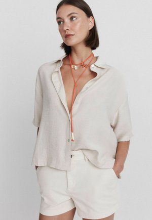 Donna che indossa una camicia beige ampia con bottoni e maniche arrotolate, pantaloncini beige e una lunga collana arancione con ciondoli a conchiglia e stella marina.