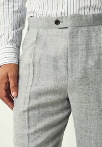 Pantalones grises ajustados con una tela texturizada, con una parte delantera plana, un solo pliegue y un cierre de botón en la cintura. Manga de camisa a rayas visible.