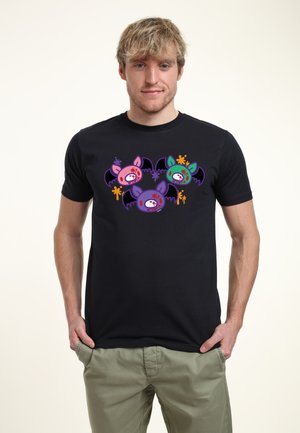 Giovane uomo con maglietta nera con disegno colorato di pipistrelli zombie in stile cartone animato, in piedi con le mani nelle tasche su sfondo semplice.