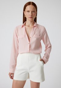 Helles rosa Langarmhemd mit Kragen, Fronttaschen und Knöpfen, kombiniert mit weißen, taillierten Shorts. Glatte, fließende Stofftexturen.