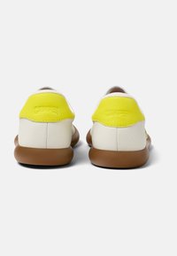 Weiße Lederschuhe mit gelbem Akzent und braunen Gummisohlen. Die Rückansicht zeigt ein abgerundetes Design und ein geprägtes Markenlogo auf der Ferse.