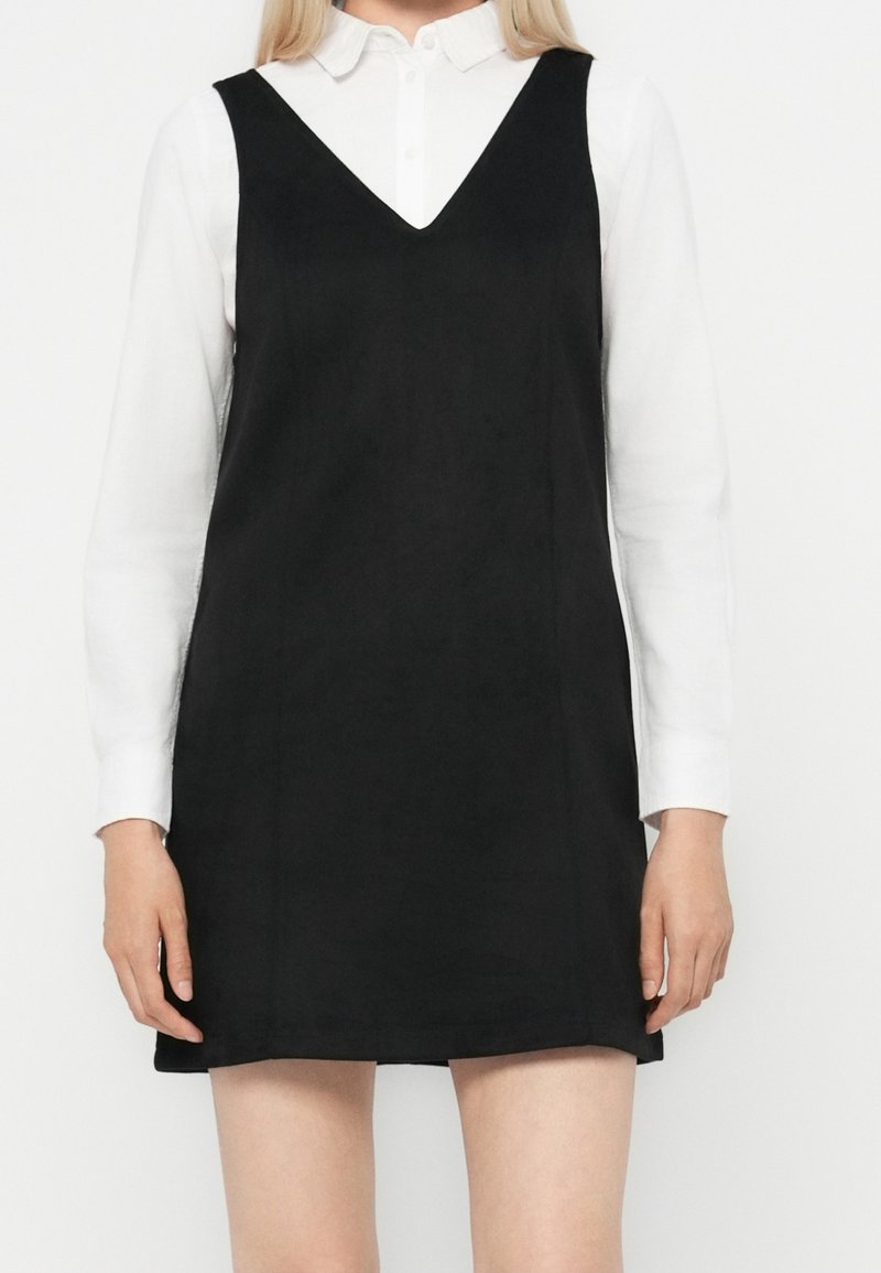Femme portant une robe noire sans manches à col en V par-dessus une chemise blanche à manches longues avec boutons, debout devant un fond blanc uni.