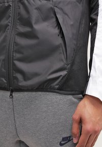 Svart zip-up jacka med strukturerad tyg och meshpaneler på sidorna, tillsammans med grå sweatpants som har en diskret logotyp.