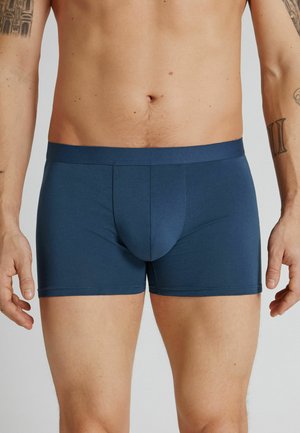 Male torso and upper legs wearing fitted navy blue boxer briefs, standing with arms relaxed at sides, visible tattoos on both arms.
