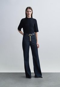 Γυναίκα που φοράει μαύρο πουλόβερ με κοντά μανίκια και σκούρο τζιν με ψηλή μέση, φαρδιά μπατζάκια, χρυσά κουμπιά μπροστά και μαύρη ζώνη.