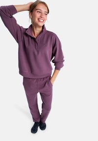 Lila sweatshirt med krage och knappar, kombinerad med matchande lila sweatpants. Båda plaggen har en mjuk textur och diskret logotyp.