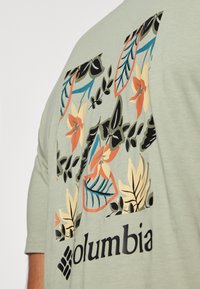 Lysegrønn kortermet t-skjorte med et blomstermønster og blader i oransje, gult, blått og svart på baksiden, med en logo i svart.