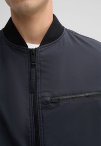 Dunkelgraue Bomberjacke mit schwarzem Kragen, Reißverschluss und einem dezenten Logo-Detail auf der Brusttasche. Glattes, leichtes Material.