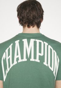 Maglietta in cotone verde con una grande stampa bianca "CHAMPION" sulla schiena, scollatura rotonda e maniche corte.
