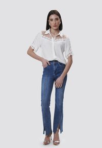Blusa bianca con dettagli in pizzo sulle maniche e sul petto, abbinata a jeans blu a vita alta con spacchi laterali e cuciture argentate.