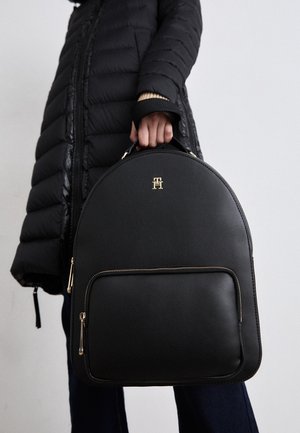 Person i sort dunjakke, der holder en sort læderrygsæk med rund form, guldlynlås og lille guldlogo på forlomma.