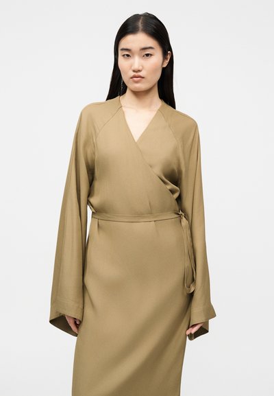 Jeune femme aux longs cheveux noirs portant une robe cache-cœur ample couleur beige avec de larges manches et une ceinture nouée, debout devant un fond clair uni.