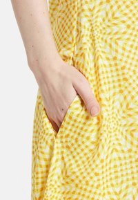 Gelbes und weißes gemustertes Kleidergewebe mit einem geometrischen Design. Eine Hand ist in einer Seitentasche platziert, wodurch die Tiefe und Struktur der Tasche zur Geltung kommt.