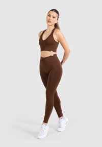 Sostenedor deportivo marrón con superficie acanalada combinado con leggings marrones de cintura alta. Presenta un diseño sin costuras y zapatillas blancas a juego.