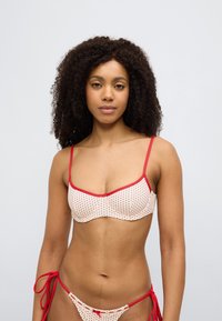 Μπικίνι τοπ με κρεμ βάση με κόκκινα πουά, κόκκινη επένδυση, ρυθμιζόμενους ιμάντες και σχεδιασμό με ειδικά διαμορφωμένα cups.