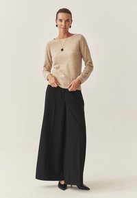 Beige trui met lange mouwen, gecombineerd met hoge zwarte wijde broek en puntige zwarte hakken. Eenvoudige hangende ketting als accent.