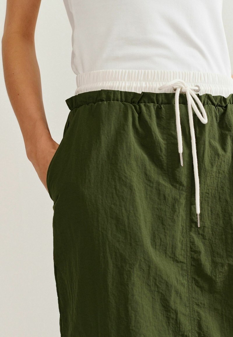 Personne portant un pantalon à cordon vert foncé avec une taille élastique blanche et des cordons blancs, main dans la poche, associé à un haut blanc.