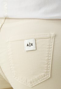 Beiga bomullsbukser med en bakficka som har en vit etikett prägad med "A|X." Slät textur och strömlinjeformat design med synlig sömnad.