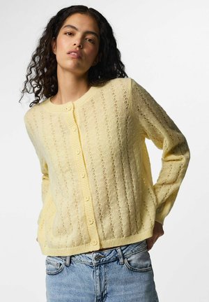 Jeune femme aux cheveux bouclés portant un cardigan en maille jaune boutonné et un jean bleu clair sur un fond uni.