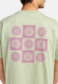 Lichtgroen katoenen T-shirt met een roze en witte zonafbeelding op de achterkant, gerangschikt in een raster met spiralen en stralen.