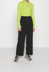 Långa svarta byxor i kombination med en räfflad, zip-up, ljusgrön topp. Skorna består av grå sneakers med flerfärgade detaljer och svarta snören.