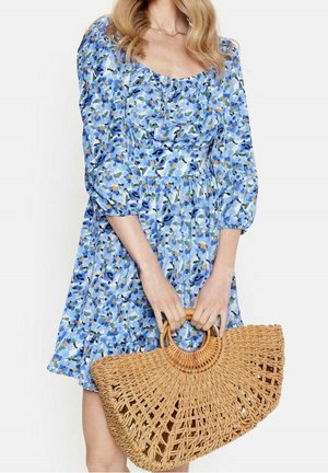 Kobieta w niebieskiej sukience w kwiaty z bufkami, trzymająca dużą plecioną słomianą torbę z okrągłymi uchwytami.