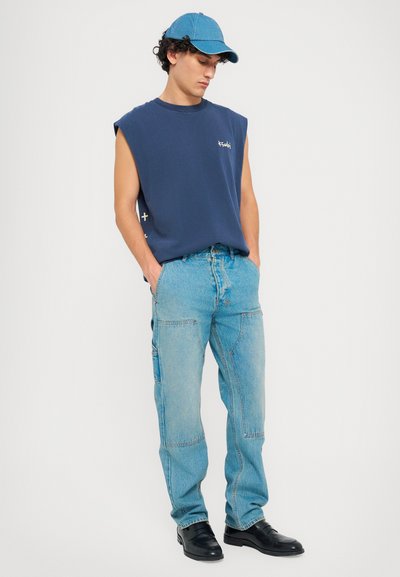 Marineblå ærmeløs sweatshirt, lys blå denimjeans med teksturerede sømme, og en matchende blå kasket. Slim-fit pasform, minimal logodetalje.
