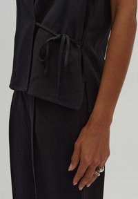 Haut noir texturé avec un nœud à la taille, présentant des fentes sur les côtés. Le tissu semble léger, associé à un bas sombre et ample.